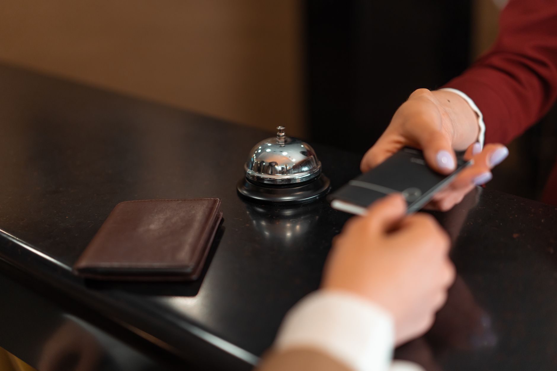 Deux mains échangent un terminal de paiement noir au-dessus d’un comptoir de réception, avec une sonnette chromée et un portefeuille en cuir.