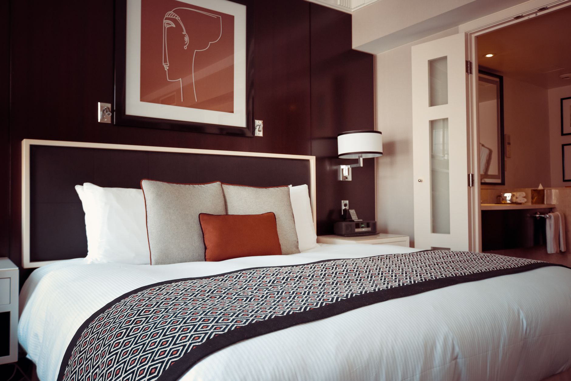 Grand lit fait avec draps blancs, chemin de lit à motifs et coussins, dans une chambre aux tons sombres avec applique et tableau.