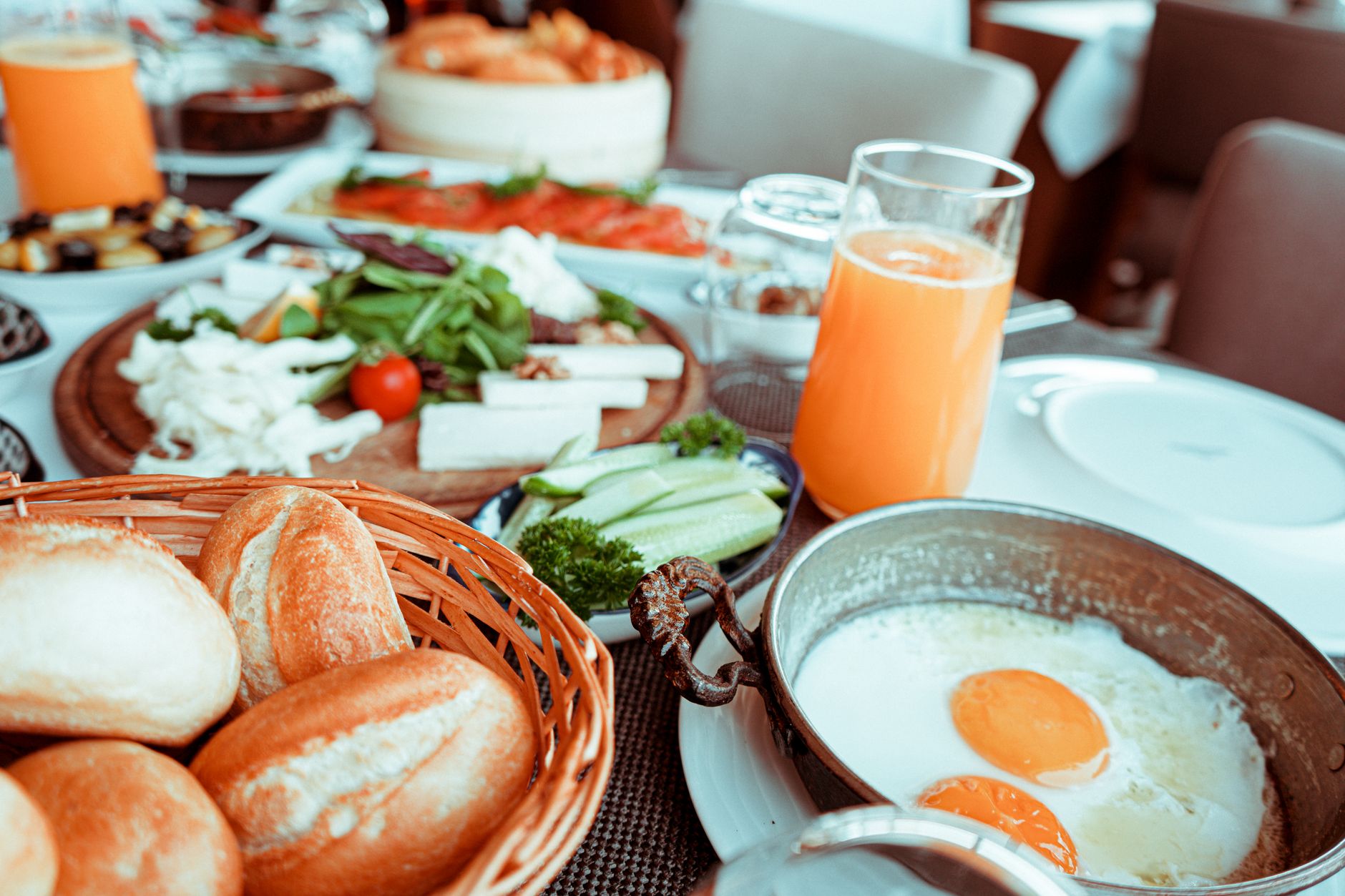 Table de petit déjeuner avec petits pains dans un panier, oeufs au plat, jus d’orange, fromage, salade et concombre.
